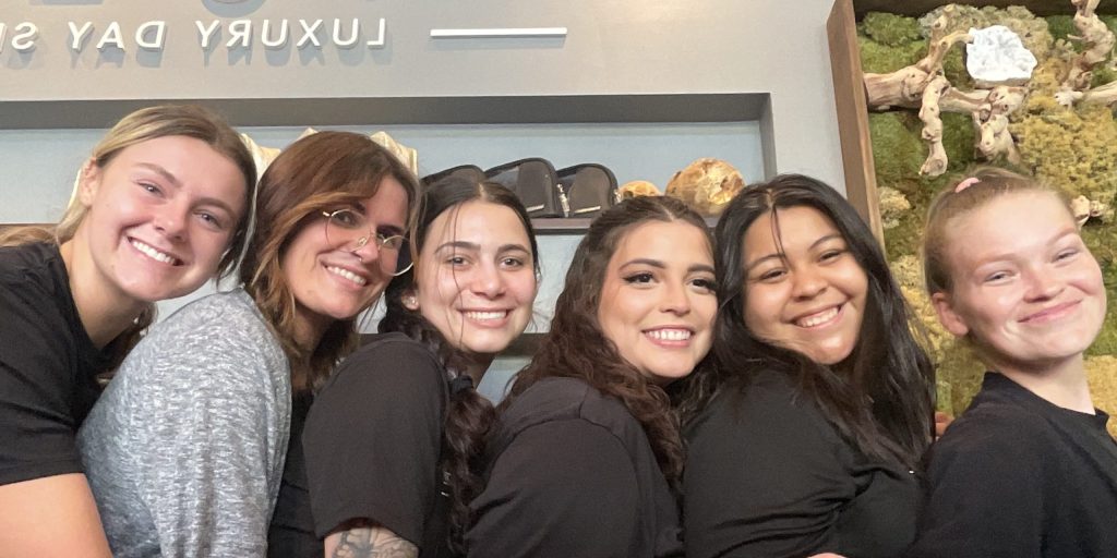 A group of six women stand close together, smiling and posing for the camera inside a spa. Behind them, a shelf with decorative items and a sign that reads "SPA" can be seen. They appear happy and relaxed in the cozy, well-lit environment.