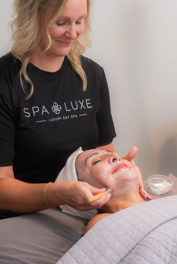 A woman with a white headband is lying down on a massage table with her eyes closed, receiving a facial treatment. An esthetician wearing a black "Spa Luxe" shirt is smiling and applying a white facial mask. A small bowl of cream is held in one hand.