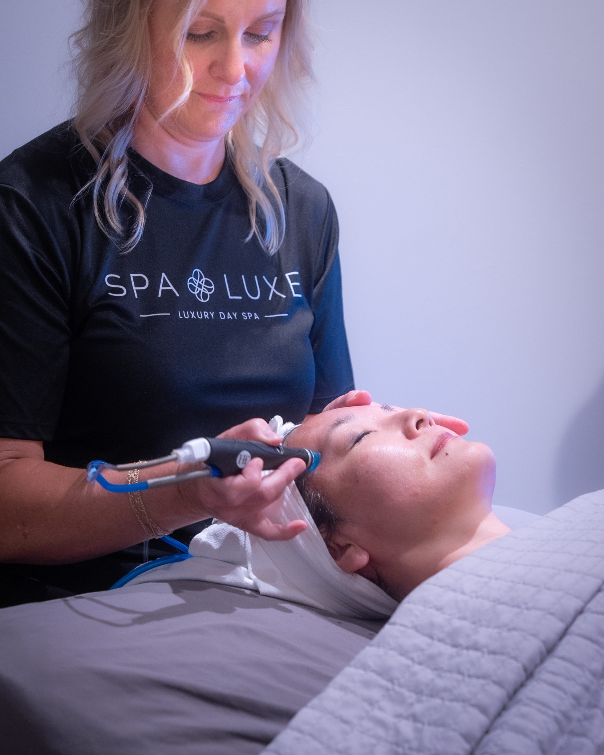 A woman performs a skincare treatment with a handheld device on another woman lying on a spa bed with a gray blanket. The woman receiving the treatment has her eyes closed and a towel wrapped around her head. The practitioner wears a black "Spa Luxe" shirt.