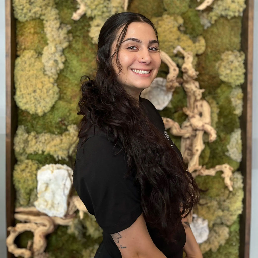 A woman with long, dark, wavy hair smiles at the camera. She is wearing a black T-shirt and has a nose piercing. Behind her is a decorative moss wall with various shades of green, white, and brown.