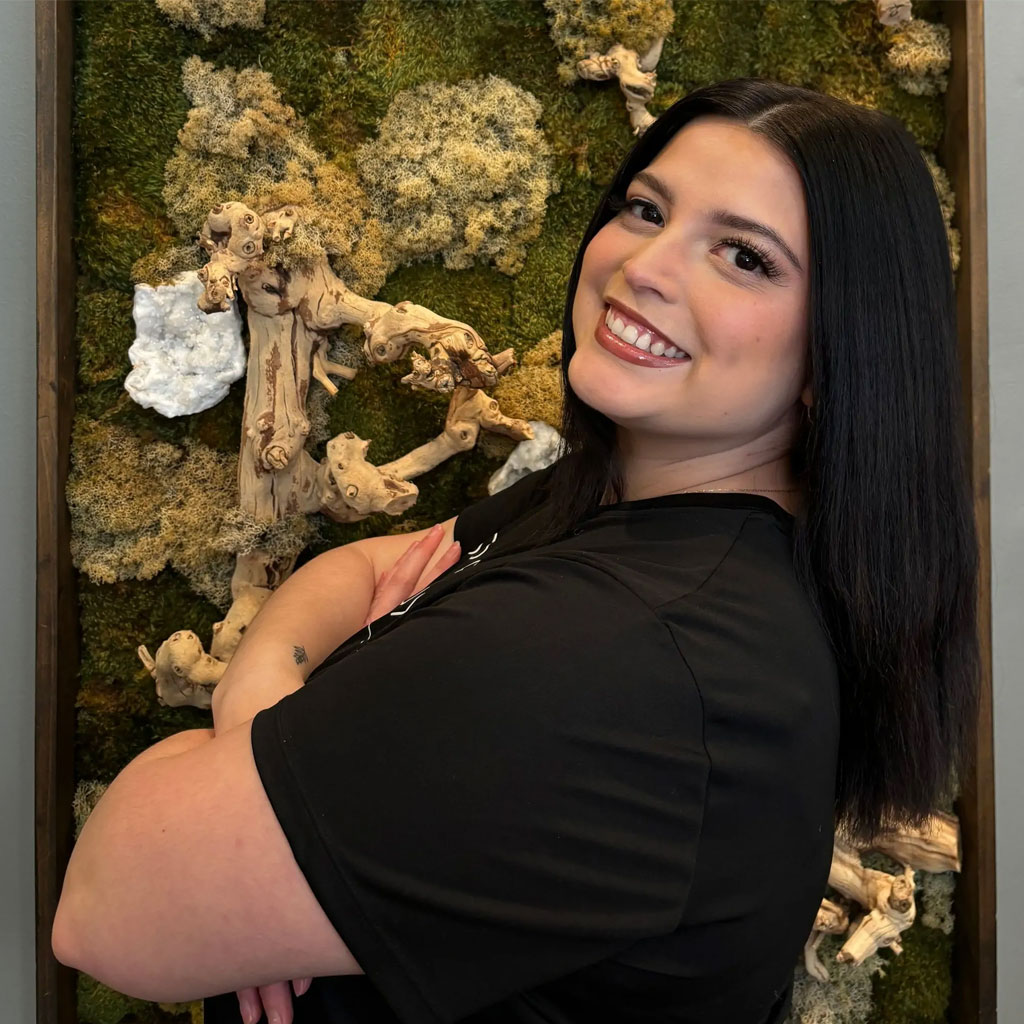 A woman with long, straight black hair stands smiling with her arms crossed. She is wearing a black T-shirt and poses in front of a textured green and white moss wall art piece. She appears confident and relaxed.