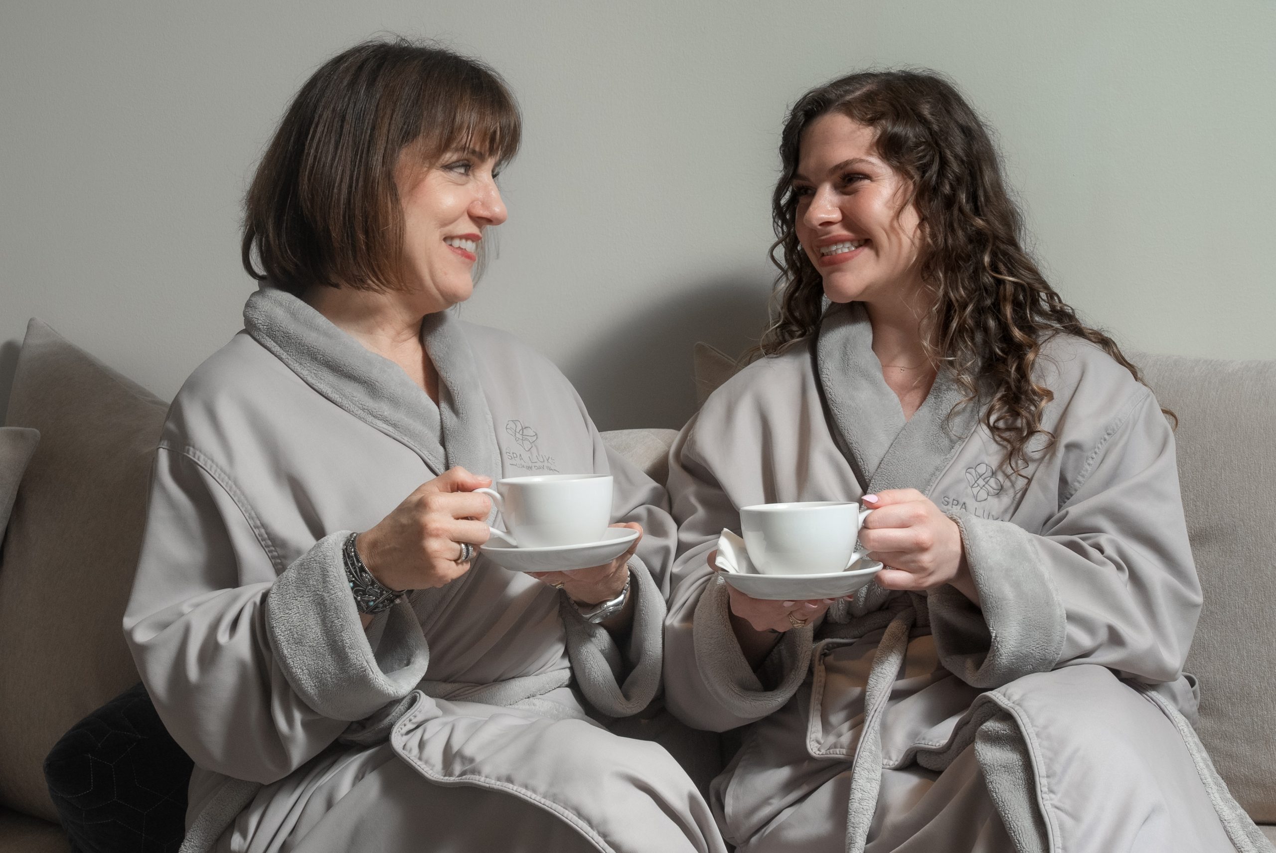 Two women sit on a couch wearing matching light grey bathrobes, each holding a white teacup and saucer. They face each other, smiling and engaged in conversation, creating a warm and friendly atmosphere.