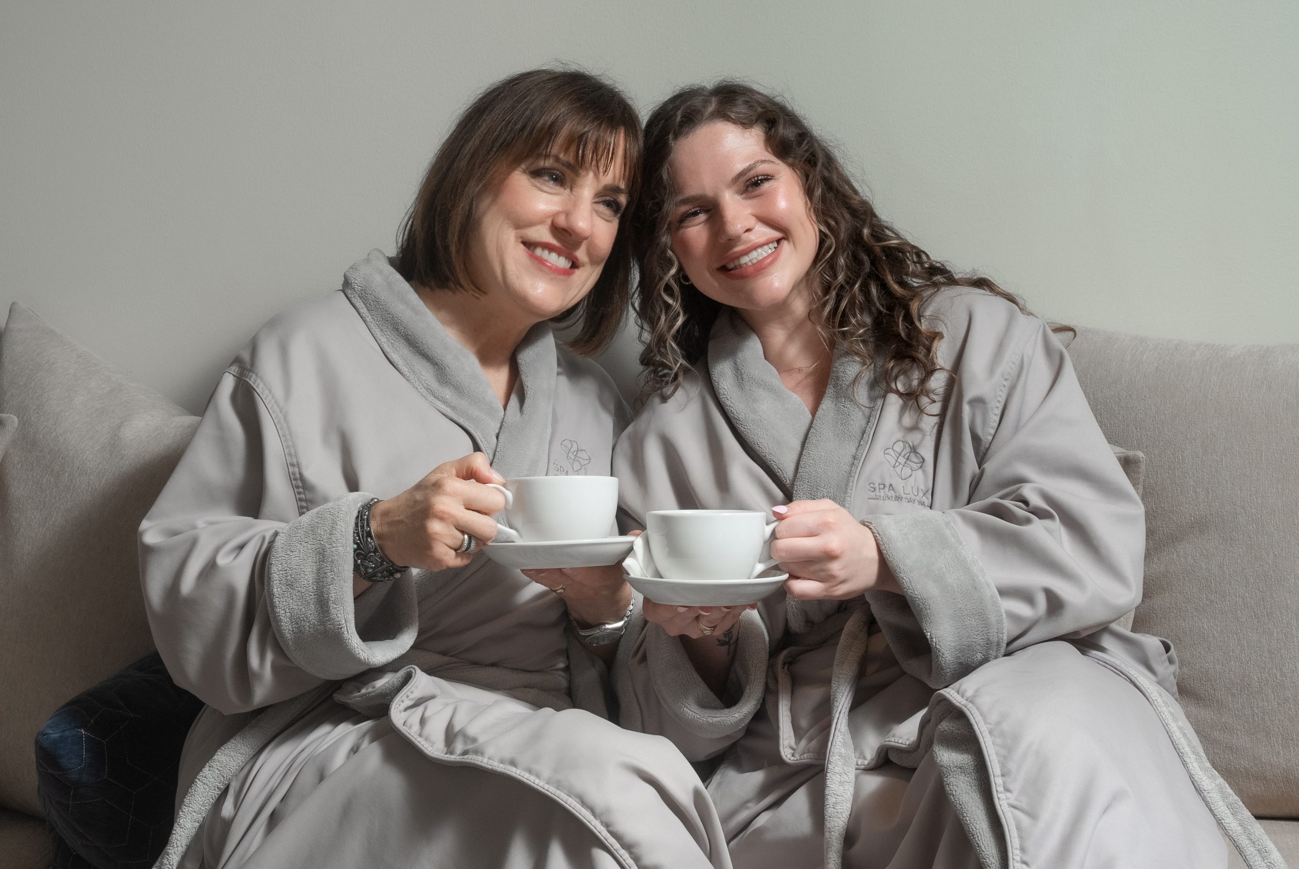 Two women, both wearing matching gray spa robes, are sitting close together on a couch. They are smiling and holding white teacups and saucers, appearing relaxed and happy. The setting looks cozy and serene, suggesting they are enjoying a moment at a spa or similar setting.