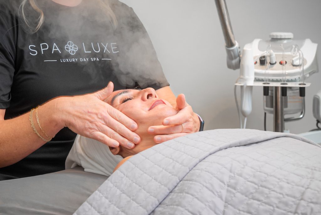 A person is receiving a facial treatment at a day spa. The individual lies on a bed with a gray blanket while a practitioner massages their face with steam emitted from a machine in the background. The practitioner's shirt reads "Spa Luxe Luxury Day Spa.