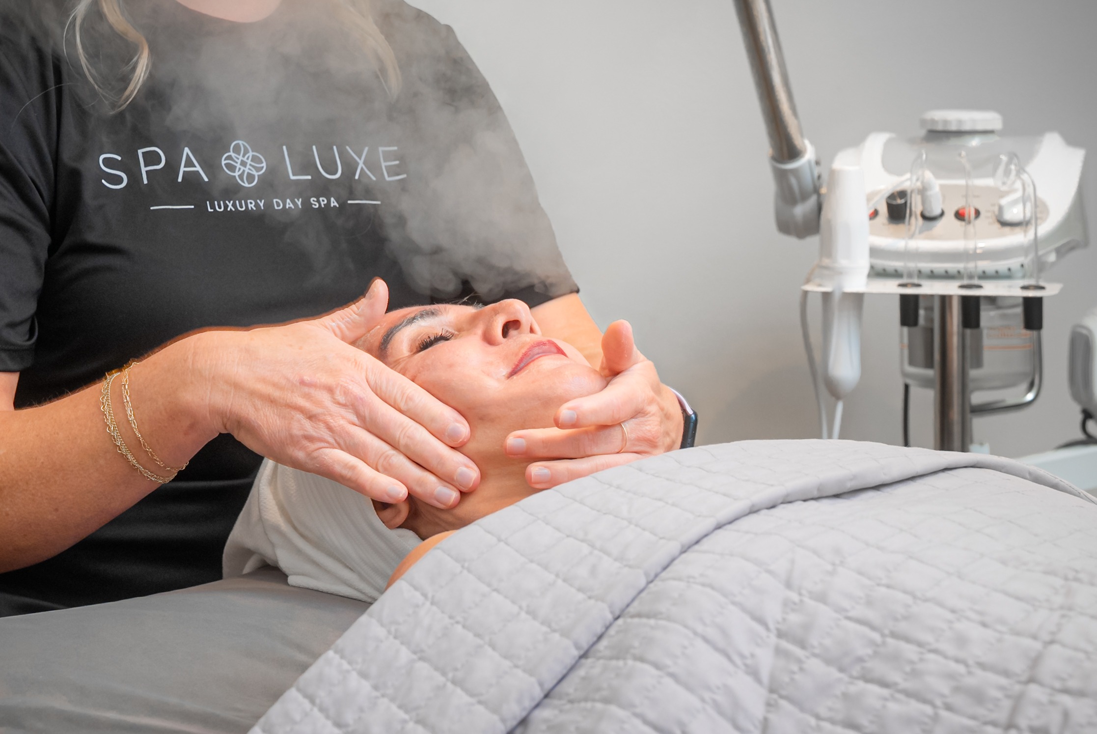 A person is receiving a facial treatment at a day spa. The individual lies on a bed with a gray blanket while a practitioner massages their face with steam emitted from a machine in the background. The practitioner's shirt reads "Spa Luxe Luxury Day Spa.