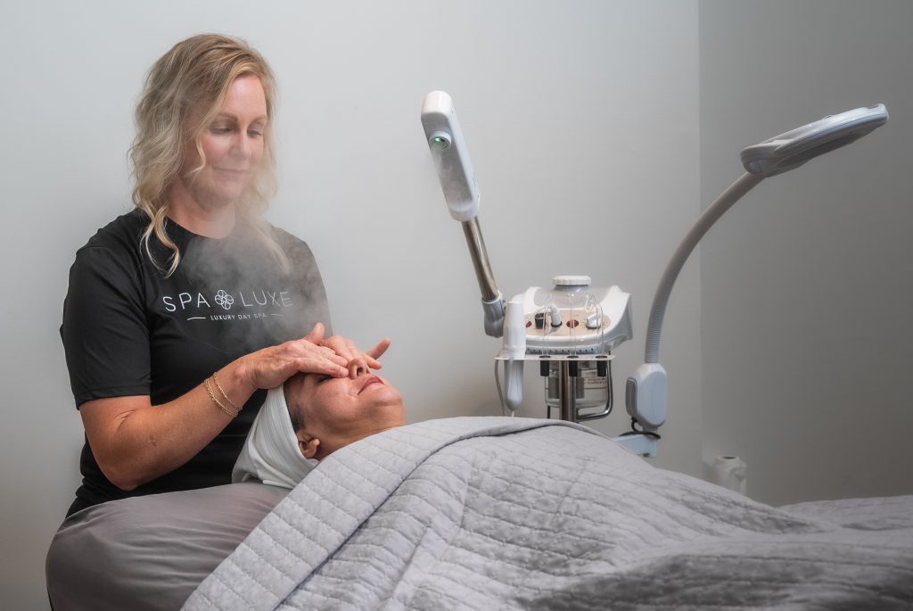 A person receiving a facial treatment while lying on a spa bed, covered with a gray blanket. A skincare specialist applies treatment to the person's face with a serene expression. A misting device and other skincare equipment are visible in the background.