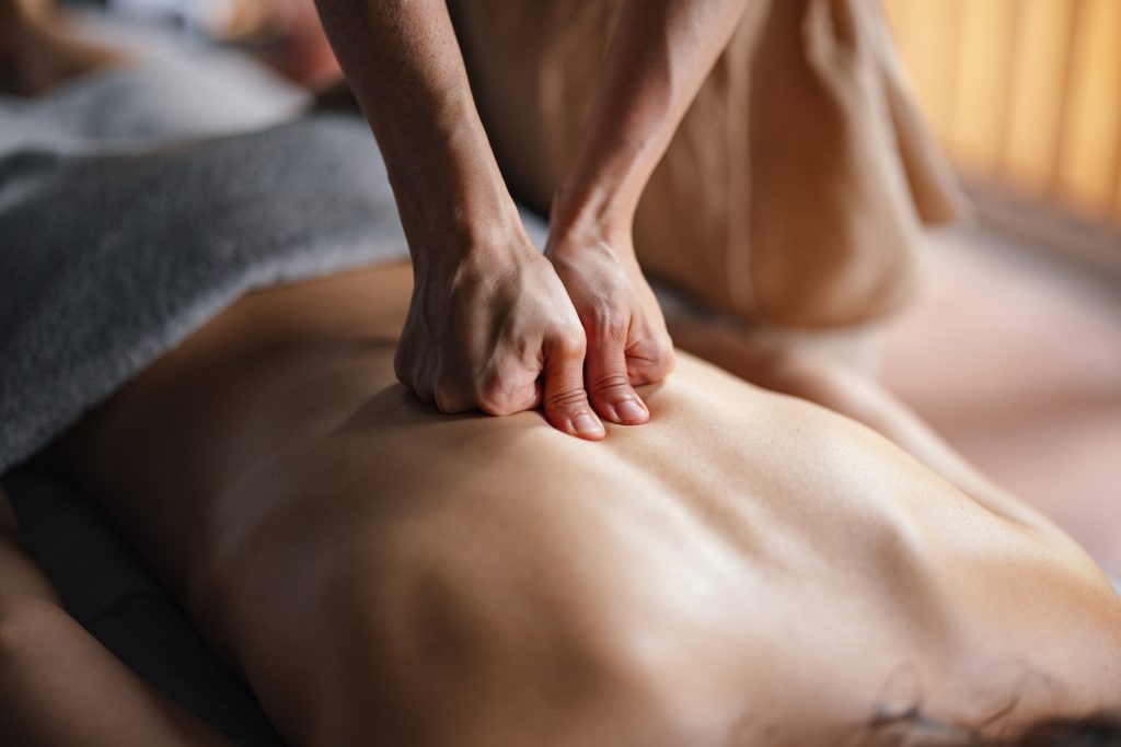 A person is receiving a deep tissue massage. The masseuse's hands are pressing firmly into the person's back, working on relieving tension and muscle knots. The person is lying down, covered partially by a grey towel. The setting appears calm and relaxing.