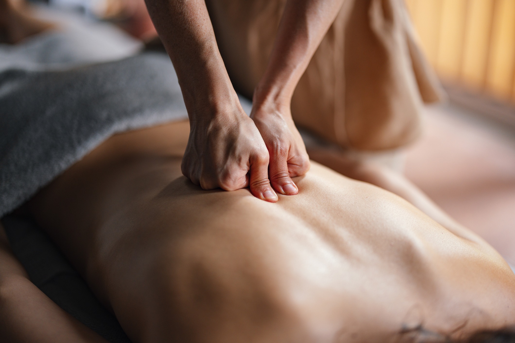 A person is receiving a deep tissue massage. The masseuse's hands are pressing firmly into the person's back, working on relieving tension and muscle knots. The person is lying down, covered partially by a grey towel. The setting appears calm and relaxing.