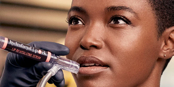 A close-up of a person receiving a skincare treatment. A gloved hand is holding a 'hydrafacial pertk Lip Revitalizing Serum' applicator near the person's lips. The person has short hair and looks upward with a serene expression.