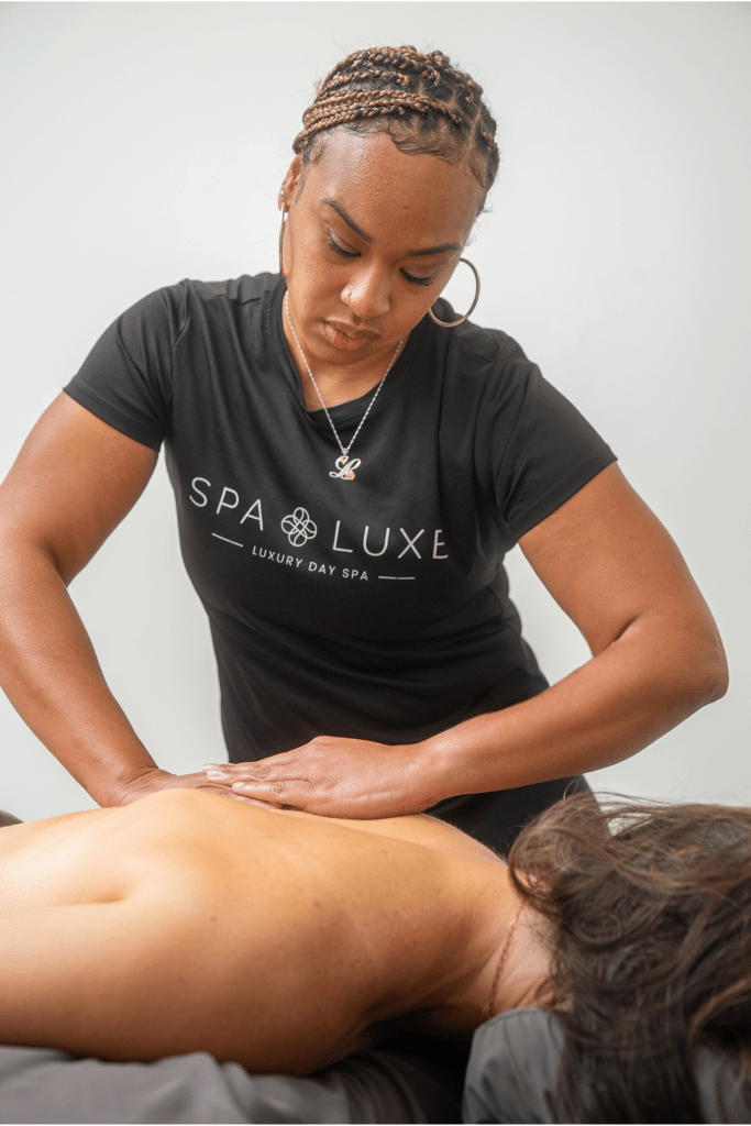 A massage therapist wearing a black "Spa Luxe" t-shirt gives a back massage to a client lying face down on a massage table. The therapist has a focused expression while working, and the background is plain and light-colored.