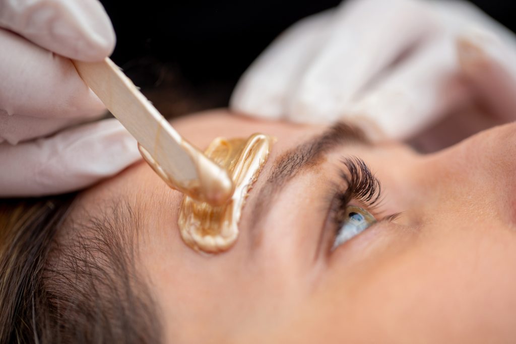 Close-up of a person receiving an eyebrow waxing treatment. A wooden spatula applies warm wax to shape the eyebrows, with a gloved hand holding the spatula. The person's eye is closed, highlighting long eyelashes and relaxed expression.