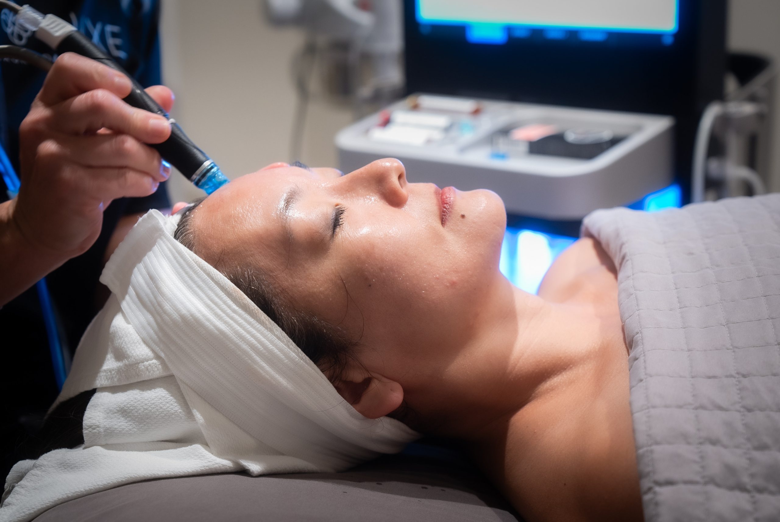 A person lies on a spa bed with eyes closed, covered with a gray blanket and a white headband. A therapist applies a treatment to their face using a handheld device. The ambiance is serene, with soft lighting and a machine visible in the background.