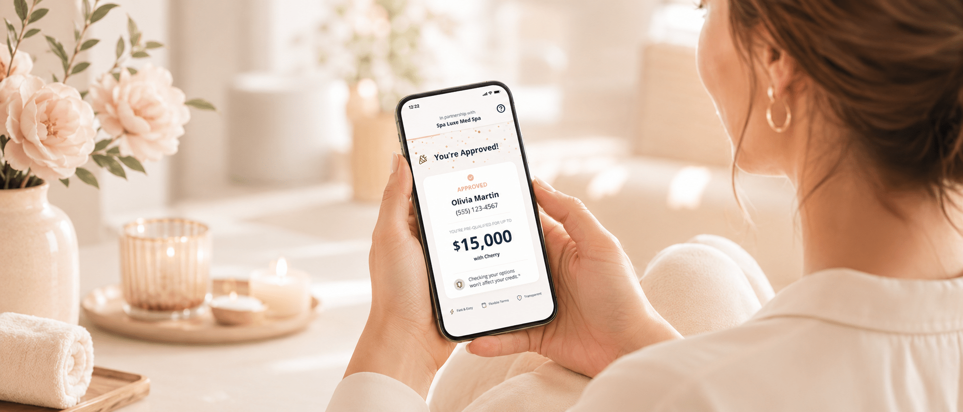 A woman sits on a sofa holding a smartphone displaying a loan approval screen, showing a $15,000 approval for Olivia Martin. The setting is cozy, with flowers, candles, and soft light in the background.