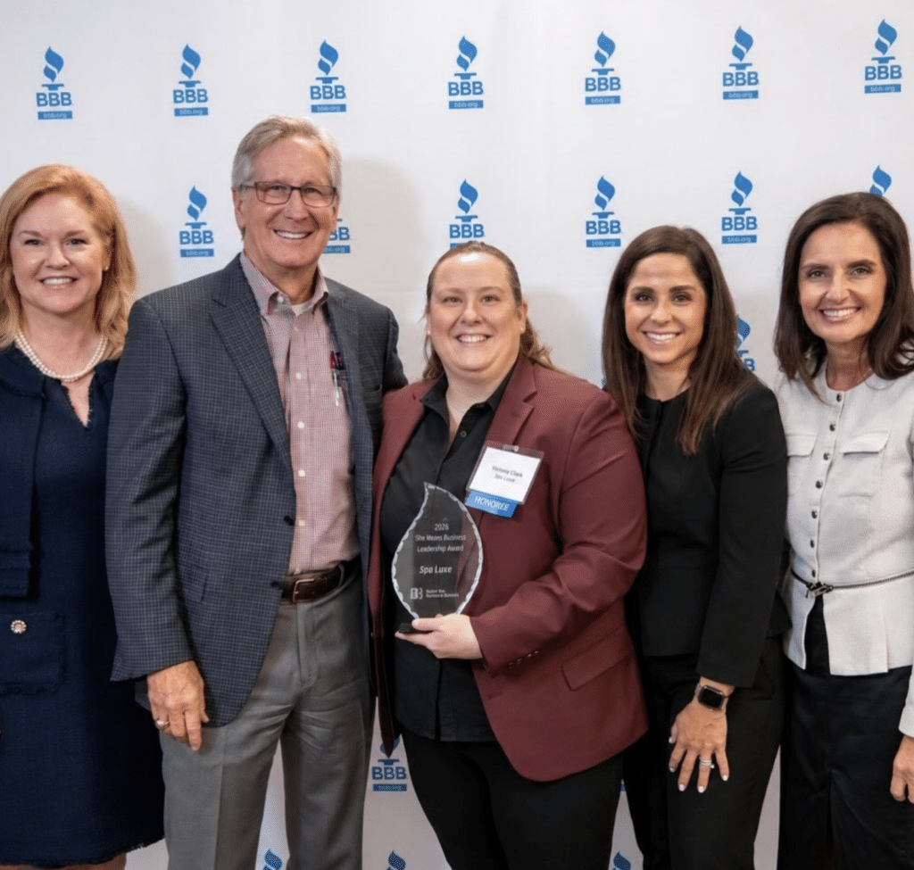 Five people stand in a row in front of a Better Business Bureau backdrop, with the center person holding an award that reads “2026 She Means Business Leadership Award, Spa Luxe.”